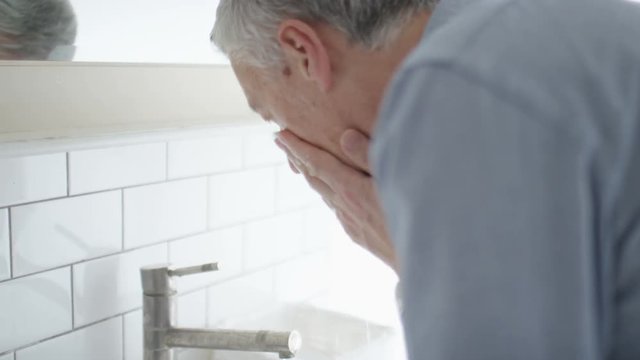 Mature caucasian male rinses his face in the sink before looking in the mirror, in slow motion