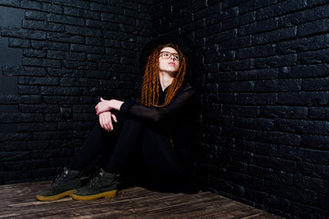 Studio shoot of girl in black with dreads, at glasses and hat on brick background.