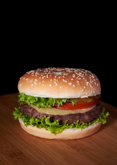 Burger on wooden surface isolated black background with copyspace on top. Hamburger includes salad, meat patty, cheese, tomato and onion. Stock photo.