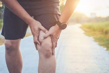 Injury from workout concept : Asian man use hands hold on his knee while running on road in the park. Shot in morning time, sunlight and warm effect with copy space for text or design - obrazy, fototapety, plakaty
