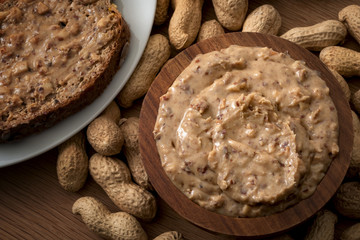 American breakfast, sweet food and snack high in calories concept with a bowl of crunchy peanut butter and a slice of bread surrounded by peanuts still in shell, isolated on wood background