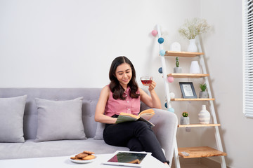 Beautiful girl at home sitting on the couch, reading book and having a coffee break