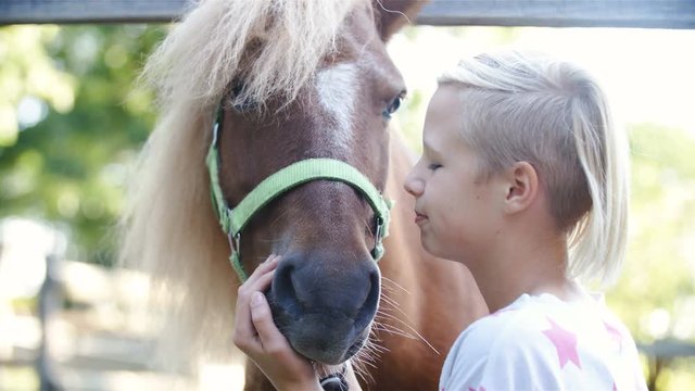 Girl kissing a pony horse with love 4K. Long shot of a cute girl in focus kissing a small brown pony with a long mane. Slow motion shot.
