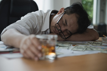 Stressed asian businessman holding a glass of whiskey. he sleep on the money, data charts, business document at office desk.  alcohol addiction - drunk businessman concept