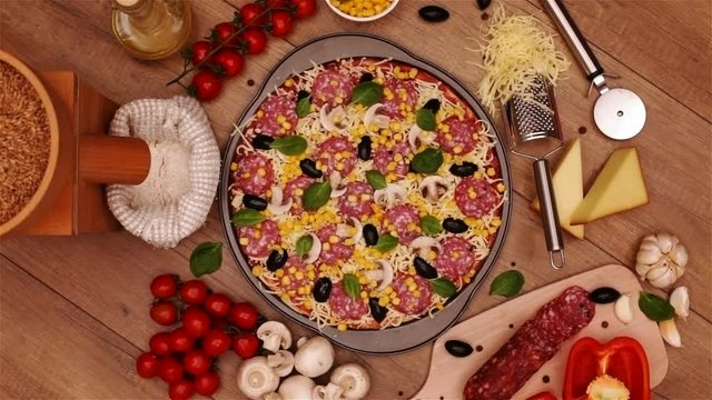 Camera spin above a homemade delicious pizza in round pan with fresh ingredients around on the table - top view