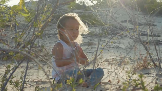 Little girl meditates at desert. Child sitting on the sand at sunset warm.
