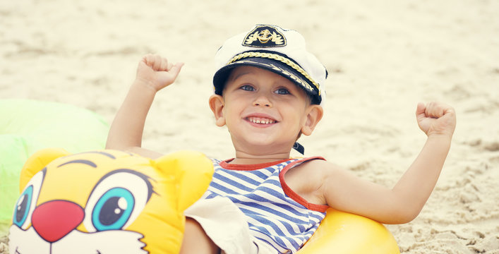 Little Caucasian boy in sailor suit and cap sitting on the beach in swim ring and laughs.