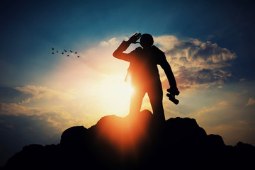 silhouette of businessman with binocular for searching a opportunity on top of mountain against sun...
