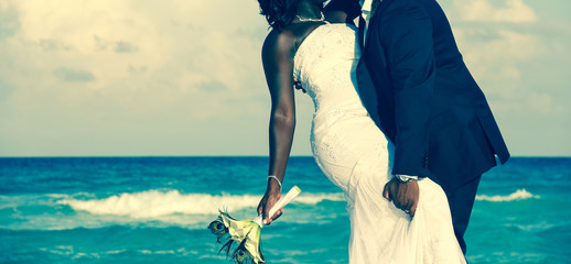Passionate Afro-American couple kissing on wedding day by the sea. Bride holding bouquet and groom grabbing her leg on the beach. Ceremony union, Valentine's day, love concept.Vintage effect