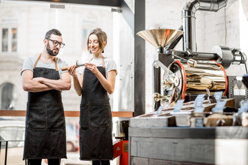 Couple of baristas in uniform checking the quality of roasted coffee beans standing near the roaster machine