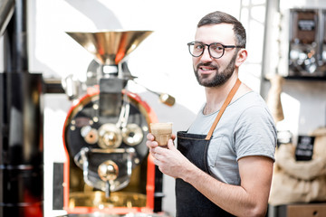 Portrait of a handsome barista in uniform standing with coffee near the roaster machine in the coffee house
