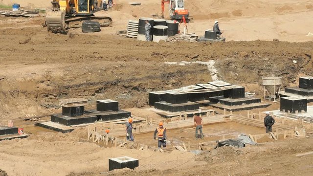 Large construction site. Excavator working on construction site. Workers in helmets and suits level surface and pour the foundation for building into wooden formwork outdoors