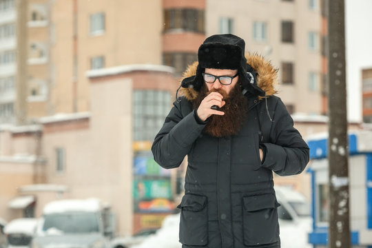 Vape bearded man in real life. Portrait of young guy with large beard in glasses and a black cap vaping an electronic cigarette in the winter.