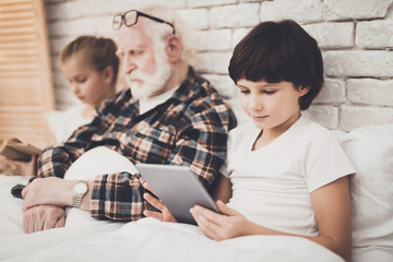 Obraz premium Grandfather, grandson and granddaughter at home. Grandpa is sleeping and children are using tablet and reading.