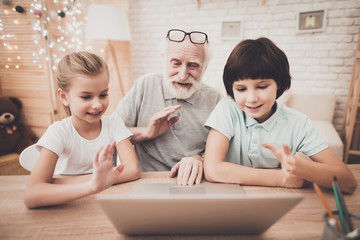 Grandfather, grandson and granddaughter at home. Grandpa and children are using skype on laptop.