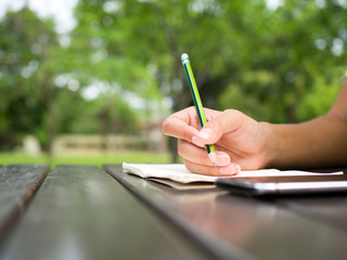 Woman hand use green pencil writing on white notebook on table in green natural park. Think out...