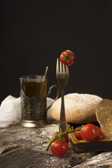 Conceptual still life of vintage fork with cherry tomato stuck on it, sliced bread and glass of tea on an ancient wooden table, and a piece of cloth