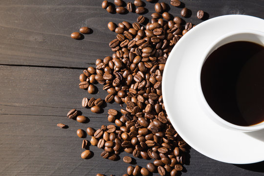 White cup of coffee with coffee beans on black wooden background and sunlight in the morning.