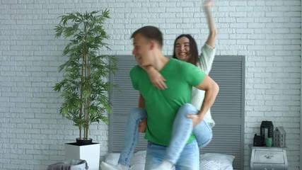 Joyful couple having piggyback ride and falling on the bed while having fun in bedroom. Excited man piggybacking his cheerful woman while beautiful couple spending leisure together in domestic room.