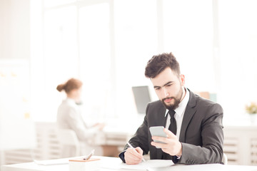Young businessman using his phone and making notes at the table at office