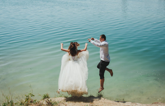 Wedding couple is jumping into the azure blue lake water. Beautiful bride in puffy dress and groom are having fun. Summer crazy emotions photo on the seaside to ocean coast. Wet wedding clothes.