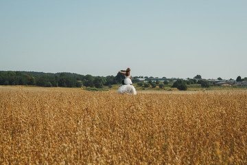 Obraz premium Wedding couple is spinning in the summer wheat field. Bride in white puffy dress hold groom's hands in black suit. Green trees and houses on the horizon. Summer heat and love story.
