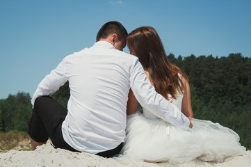 Wedding couple is sitting and hugging on the white sandy lake beach. Romantic hug and embrace of bride and groom in summer heat love story. Heating sun and weather. Travel lifestyle photo.