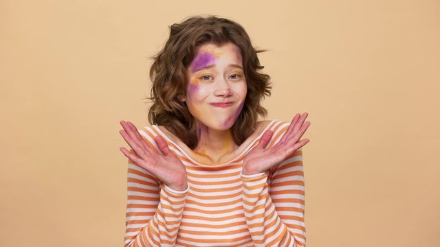 Portrait of brunette woman having multicolored painted spots on face thrugging and throwing up hands in unawareness, isolated over beige background. Concept of emotions