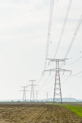View of a row of steel lattice transmission towers supporting a high-voltage overhead power line across the french countryside, passing over plowed land under a pale light.