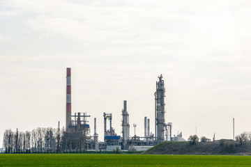 View of an oil refinery with smokestacks surrounded by crop fields in the french countryside under a pale sunlight.
