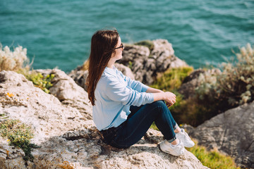 Naklejka premium Young girl stays on the stones facing the sea. The country relaxes and meditates at the spring sun in the city