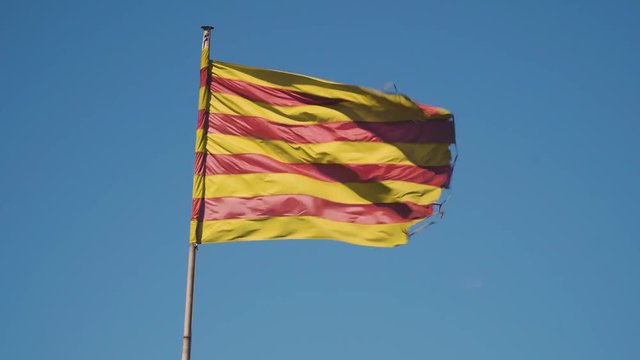 The official flag of Catalonia, Spain, is waving heavily due to a strong wind. The colors of the striped flag are yellow and red.