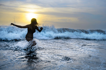 Fototapeta na wymiar silhouette of young woman running happy on sea water playing with big waves free and joyful splashing on Summer sunset beach
