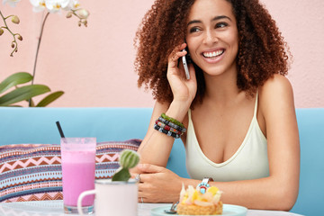 Positive dark skinned young cute African American female has pleasant talk via modern smart phone, recreats in coffee shop, drinks smoothie and eats cake, enjoys leisure time and communication