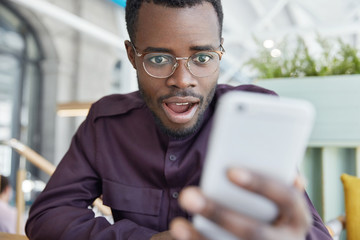 Shocked dark skinned businessman in eyewear, recieves notification on modern smart phone, gets notice to pay bills, has surprised expression. African American male reads shocking news in internet.