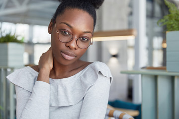 Beautiful dark skinned clever female student in big round eyewear, feels tired after preparing for exams, looks confidently into camera. People, ethnicity, facial expressions and lifestyle concept