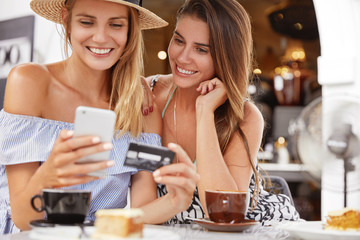 Portrait of female models make shopping online, use smart phone, credit card, sit together at cafe interior with aromatic coffee, have positive looks. Best friends recreat together, uses technologies