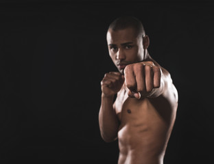 Waist up portrait of african sporty guy with naked body who looking calmly at camera and beating left hand in front. Focus on active fist. Isolated on black background and copy space in left side