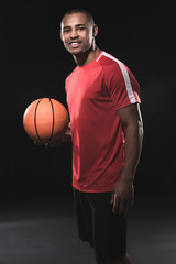 Full length portrait of young african guy keeping basketball while standing in half-turnover and looking at camera with happily smile. Isolated
