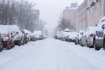 Snowfall in the empty street. Rows of parked cars covered with snow. Buildings and park can be seen through spots of falling snowflakes. White winter in a city