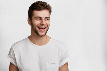 People, happiness and emotions concept. Smiling joyful young male with attractive look, dressed in casual white t shirt, looks gladfully aside, poses against studio background with copy space