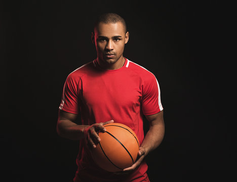 Waist up portrait of african guy with basketball in hands. Calm fearless man looking at camera with desire to victory. He is always ready to play. Isolated on black background