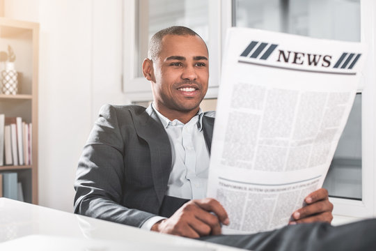 Portrait of young african guy reading newspaper. Man in respectable suit sitting near table and smiling