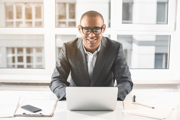 Waist up portrait of african businessman working laptop. He is smiling happily at camera. Documents and mobile phone are on table. Wide window overlooking the neighboring building is in the background