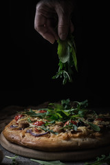 Fresh pizza with tomatoes, cheese, chicken and mushrooms. Italian pizza sprinkled arugula, tired of labor by hands. Black rustic background. Closeup