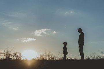 Fototapeta premium Side view profile of adult and little male silhouette standing on grass opposite each other against blue sky background. Copy space in left side