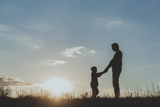 Side view profile of adult and child male silhouette standing on grass opposite each other and holding hands against sunset sky background. Copy space in left side