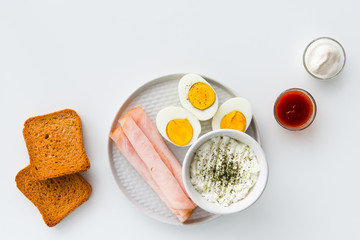 Breakfast, half boiled eggs, ham, fresh cheese sprinkled with dill, toast, horseradish sauce and sweet and sour sauce. Flat lay on white background.