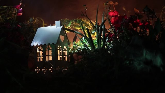 Slider shot. Fantasy night decoration. Small beautiful house in grass with light. Old house in forest at night with moon. Dark foggy lighted background. Selective focus