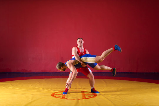 Two strong wrestlers in blue and red wrestling tights are wrestlng and making a  making a hip throw  on a yellow wrestling carpet in the gym. Young man doing grapple.